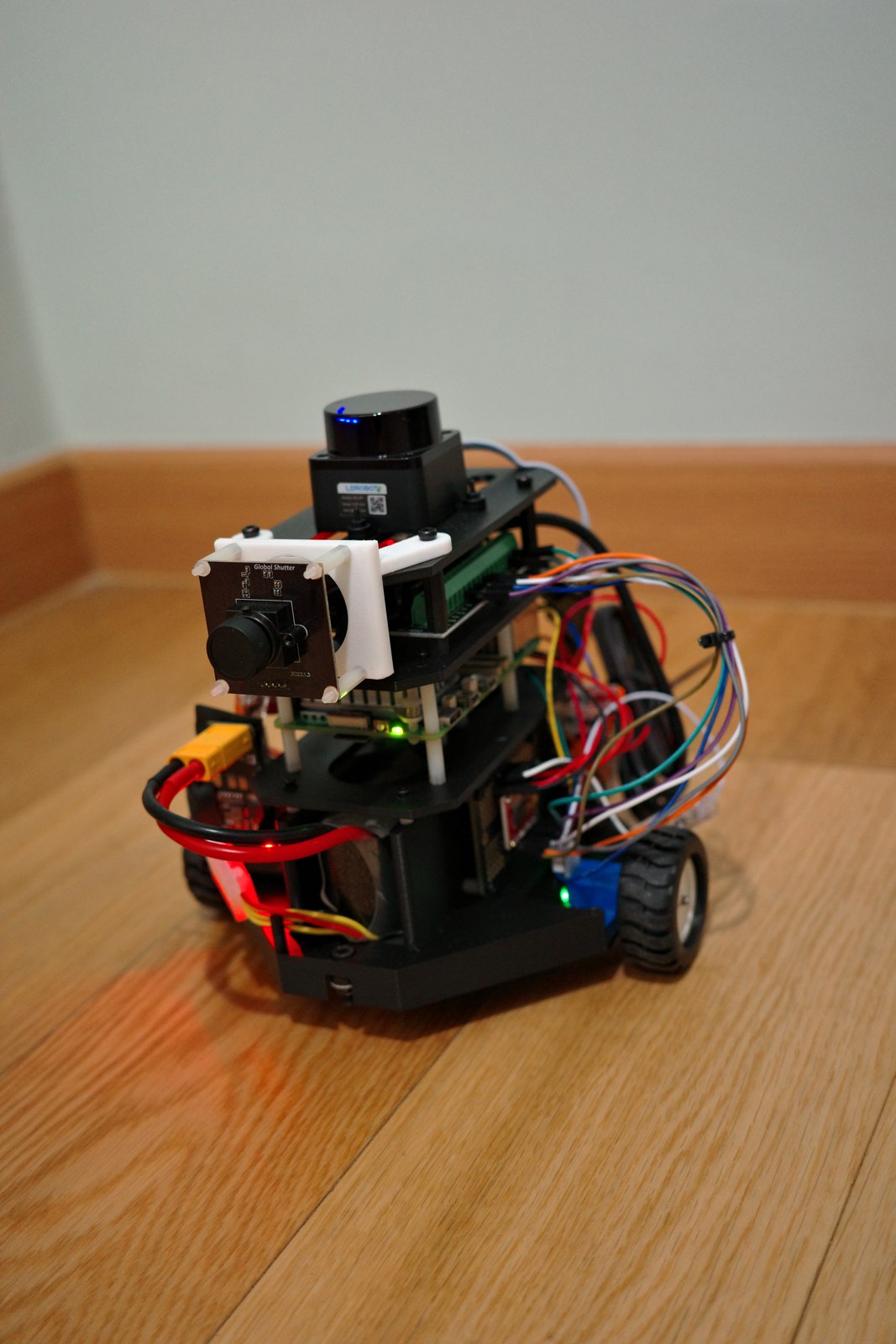 The Lab Scout rover on a wood floor: LiDAR on top, a global-shutter camera below it, stacked boards and wiring, two small wheels on a 3D-printed chassis